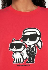 Rajzfilm egy öltönyös, lófarkas személyről napszemüvegben, aki egy piros pólót viselő, szintén napszemüveget viselő bolyhos macska mellett áll.