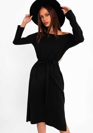 Kobieta z długimi brązowymi włosami, ubrana w czarną sukienkę odsłaniającą ramiona oraz szerokim rondem czarny kapelusz, która poprawia kapelusz obiema rękami.