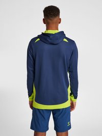 Marinblå fukttransporterande hoodie med ljusgröna detaljer, featuring en huva, långa ärmar och en strukturerad tyg. Matchad med blåa shorts.