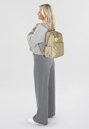 Tansfarbener Nylon-Rucksack mit zwei Fronttaschen, Neon-Grünen Akzenten, Reißverschlüssen und verstellbaren Trägern, die lässig über einer Schulter getragen wird.