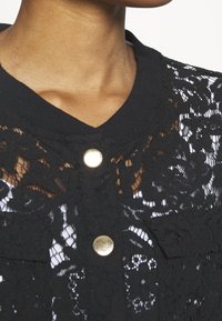 Musta pitsimekko, jossa on pyöreä pääntie, kultaiset napit ja monimutkaiset kukkakuviot, pehmeä kangas tekstuuri alla.