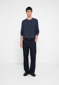 Πλεκτό πουλόβερ ναυτικού μπλε με σχέδιο καλωδίου, σε συνδυασμό με σκούρο παντελόνι. Το μοντέλο στέκεται μπροστά σε απλό λευκό φόντο, φοράει μαύρα παπούτσια.