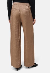 Braune, weit geschnittene Hose mit strukturierter, glänzender Oberfläche, ausgestattet mit einem elastischen Bund und Gesäßtaschen, kombiniert mit schwarzen Schuhen.