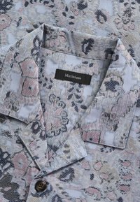 Chemise bleu pastel avec un motif floral abstrait en rose, beige et noir, dotée d'un col pointu et d'un bouton noir.