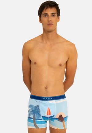 Homme debout, les mains derrière le dos, portant un boxer coloré avec un motif de palmier, de voilier et de scène de plage au coucher du soleil.