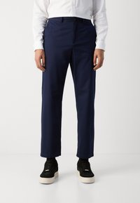 Marineblå bukser laget av glatt stoff, med rett passform, sidelommer og knappestengning. Kombinert med svarte joggesko.