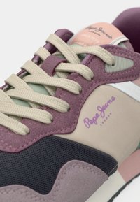 Multi-colored sneaker featuring shades of purple, green, and beige, with textured fabric, prominent logo, and woven laces.