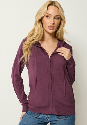 Blond vrouw met een paarse hoodie met ritssluiting en blauwe spijkerbroek, staand tegen een effen achtergrond, kijkt naar de zijkant.