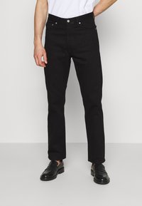 Jeans de mezclilla negros con corte recto, diseño de cinco bolsillos y detalles metálicos. Llevados con una camiseta blanca y mocasines negros.