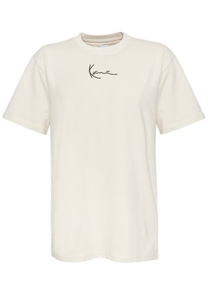 Camiseta de algodón color crema, con mangas cortas, cuello redondo y un pequeño logotipo negro de firma en el lado izquierdo del pecho. Textura suave, corte regular.