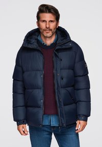 Granatowa kurtka puchowa z kapturem, z pikowanym wzorem, z przodu zapinana na guziki oraz suwak. Nosi się ją na teksturowanym burgundowym swetrze.