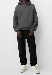 Felpa con cappuccio grigio antracite dalla vestibilità oversize, con polsini e orlo ribattuti, abbinata a pantaloni jogger neri e sneaker crema. Tessuto liscio.