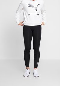 Persona che indossa una felpa bianca Puma, leggings neri con logo sulla caviglia e sneakers bianche Puma, in piedi contro uno sfondo semplice.