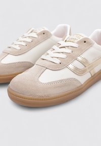 Zapatillas deportivas de ante beige y piel blanca con cierre de cordones, detalles en dorado y suelas de goma. Acabado texturizado y diseño clásico.