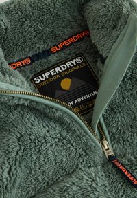 Flauschige grüne Fleece-Jacke mit Reißverschluss, goldfarbener Reißverschluss und schwarzem Patch mit einem Sonnen- und Bergdesign; "SUPERDRY"-Branding in Orange.