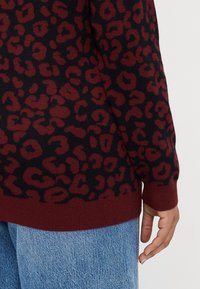 Suéter de padrão de leopardo em vermelho e preto com uma barra canelada em bordo, feito de material tricotado suave, apresentando mangas longas e um corte descontraído.