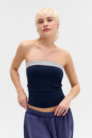 Ung person med kort blondt hår iført en navyblå tube top med grå kant og løse lilla bukser, poserer mod en hvid baggrund.