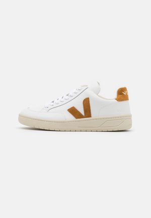 Baskets basses blanches avec semelle beige, logo V en daim orange sur le côté, patte arrière orange assortie et lacets blancs sur fond uni.