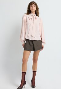 Blusa rosa chiaro con un grande fiocco e maniche lunghe a sbuffo, abbinata a una gonna corta marrone e calze al ginocchio bordeaux.