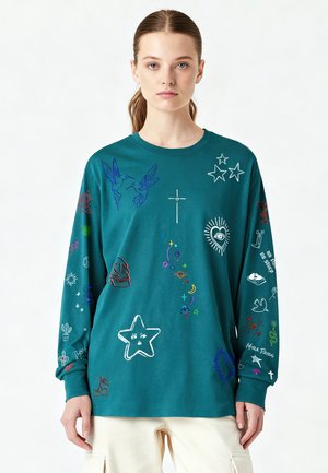 Vrouw met een teal kleurige lange mouwen shirt met kleurrijke geborduurde symbolen zoals sterren, vogels, schedel, hart, kruisen en planten, gecombineerd met een lichte broek.