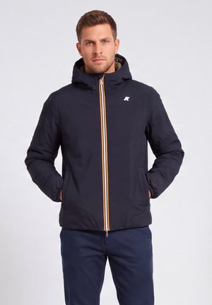 Uomo che indossa una giacca nera con cappuccio con dettagli arancioni e bianchi sulla cerniera, mani nelle tasche, in piedi davanti a uno sfondo chiaro e uniforme.