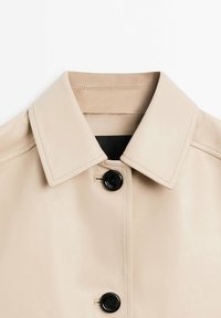 Beige lædertrenchcoat med en klassisk krave og to sorte knapper. Glat tekstur med synlige sømdetaljer.