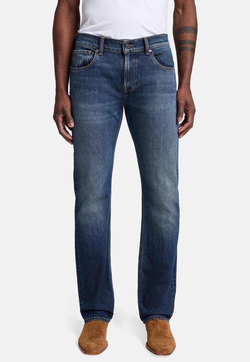 Jeans in denim di un blu scuro con una leggera sbiaditura; taglio dritto; design a cinque tasche; chiusura con bottone e cerniera in metallo; indossati con scarpe color sabbia.