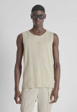 Jonge man met oversized zwarte zonnebril, mouwloze beige top en licht beige broek, staand tegen een effen lichtgrijze achtergrond.