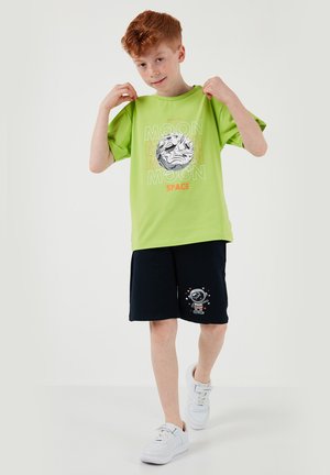 Garçon aux cheveux roux portant un t-shirt vert citron avec un motif de lune et un short bleu marine avec une illustration d'astronaute, souriant et tenant le col de son t-shirt.