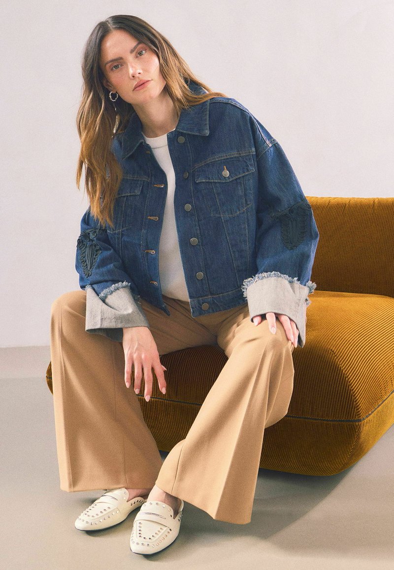 Giacca di jeans blu con polsini grigi sfrangiati, bottoni a pressione e dettagli ricamati, abbinata a pantaloni beige a gamba larga e scarpe slip-on bianche.