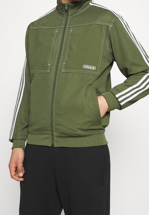 Mand iført olivengrøn Adidas-jakke med hvide striber på ærmerne, lynlås foran og logo på brystet, kombineret med sorte bukser.