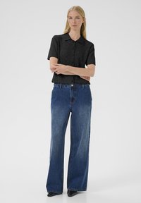 Femme blonde debout, les bras croisés, portant un haut noir à boutons à manches courtes, un jean large bleu et des chaussures noires à bouts pointus.