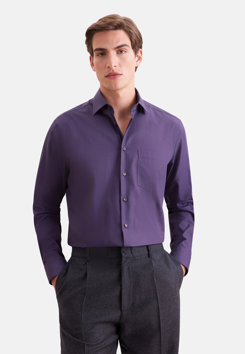 Jeune homme aux cheveux courts bruns portant une chemise violette ajustée à manches longues boutonnée et un pantalon gris foncé, mains dans les poches, fond neutre.