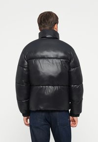 Chaqueta de plumas negra con cuello alto, acabado brillante, patrón acolchado horizontal y puños elásticos, combinada con pantalones de mezclilla.