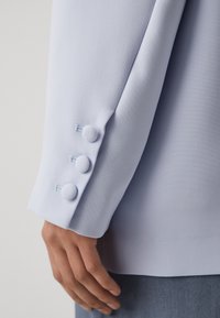 Svetle modrý blejzr s rukávem vyrobeným z texturované látky, třemi ozdobnými knoflíky a čistými liniemi. Manžeta je mírně ohrnutá.