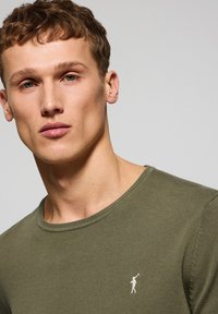 Suéter ajustado de cuello redondo en verde oliva, fabricado con suave material de punto, que presenta un pequeño logo blanco en el lado izquierdo del pecho y un cuello acanalado.