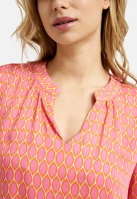 Kurzarmbluse mit V-Ausschnitt, aus leichtem Stoff. Sie hat ein ovales Muster in Rosa und Orange mit leichten Falten.