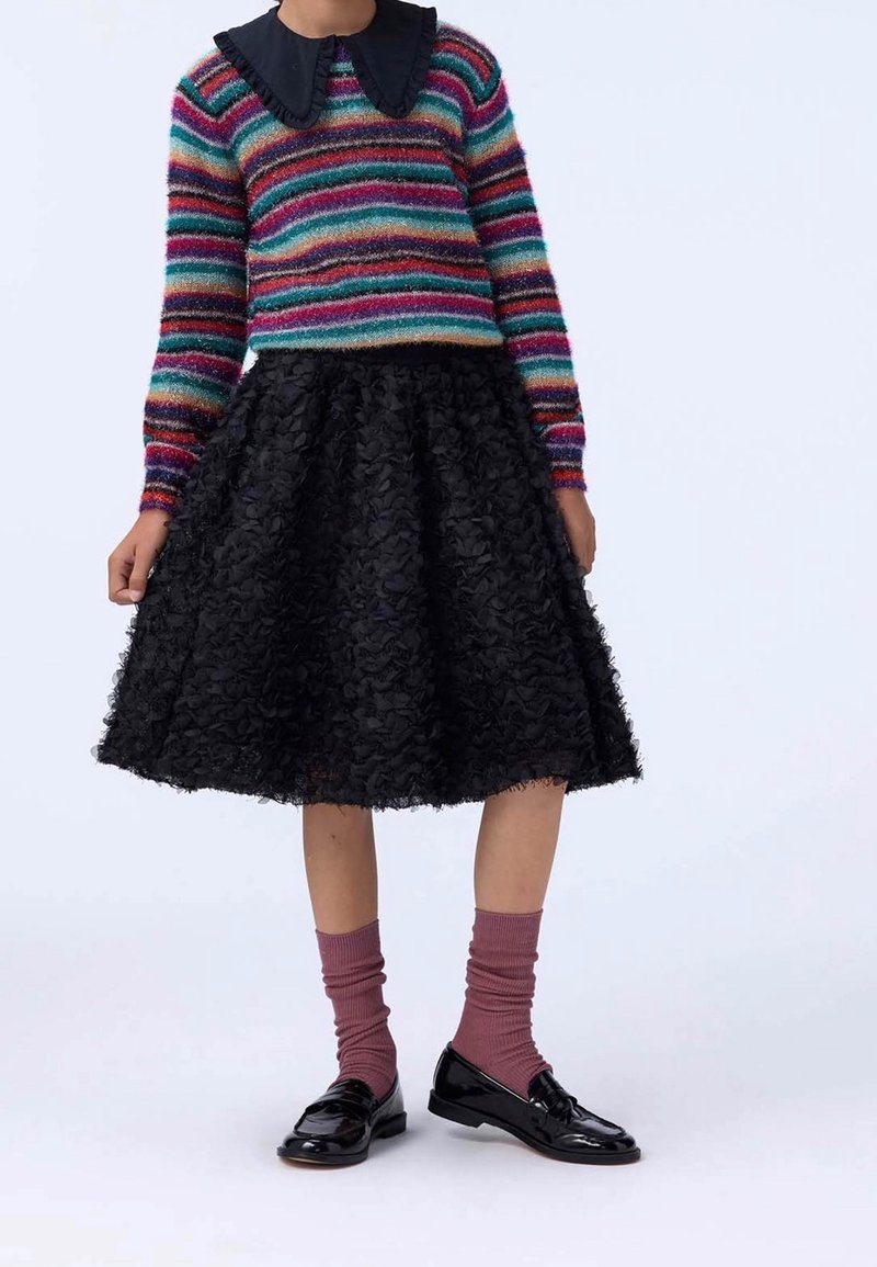 Personne portant un pull rayé multicolore avec un col noir froufroutant, une jupe noire texturée longueur genoux, des chaussettes violettes et des mocassins noirs brillants.