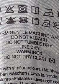 Metka pielęgnacyjna z instrukcjami prania w czarnym tekście na jasnoszarym tle tkaniny, zawierająca symbole pralnicze oraz różne wytyczne dotyczące pielęgnacji.