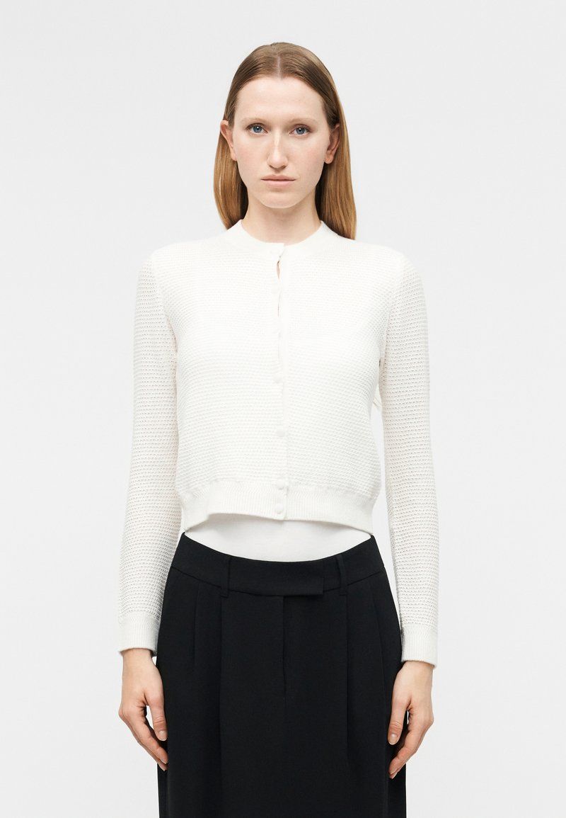 Γυναίκα με ίσια μαλλιά φοράει λευκό υφασμάτινο πουκάμισο με κουμπιά και ψηλόμεση μαύρη φούστα, στέκεται μπροστά σε απλή λευκή φόντο.