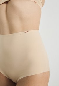 High-waisted nude panties made of a smooth fabric; features a seamless design with a small metallic logo detail at the waist.