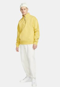 Felpa gialla Nike con mezza zip, maniche lunghe, polsini e orlo a coste; abbinata a pantaloni bianchi e sneakers bianche. Tessuto morbido.