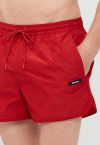 Sarkanas peldšortas, izgatavotas no viegla auduma. Iezīmējas ar elastīgu jostu ar auklu un sānu kabatu. Redzama melna Calvin Klein birka.