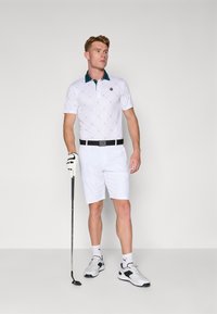 Hvid golf polo med grøn krave, diamantmønster, parret med hvide shorts og sort bælte. Hvide sportsko og golfkølle inkluderet.