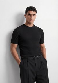 Άνδρας που φοράει μαύρο ριμπ κοντομάνικο πουκάμισο και μαύρο παντελόνι με ζώνη, στέκεται μπροστά σε έναν απλό λευκό τοίχο με τα χέρια στις τσέπες.