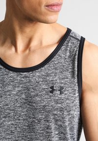 Primer plano de una persona que lleva una camiseta sin mangas atlética gris y negra con un logo de Under Armour en el pecho.