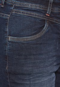 Jean en denim bleu marine avec un design classique à cinq poches, coutures contrastées et détail de poche courbé. Texture lisse, coupe slim.