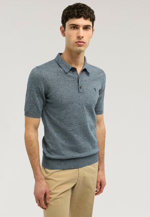 Młody mężczyzna w niebieskiej koszulce polo z krótkim rękawem i beżowych spodniach, stojący na tle jednolitego jasnoszarego.