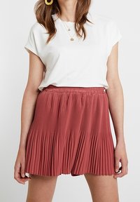 Pleated, rust-colored skirt with an elastic waistband, paired with a white short-sleeve top. Silhouette is casual and flowing.