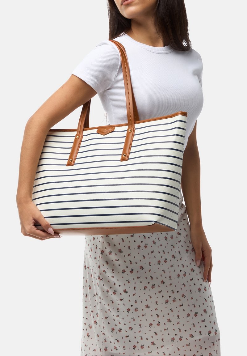 Femme tenant un grand sac cabas blanc avec des rayures horizontales bleu marine et des anses en cuir marron, portant un haut blanc et une jupe fleurie.